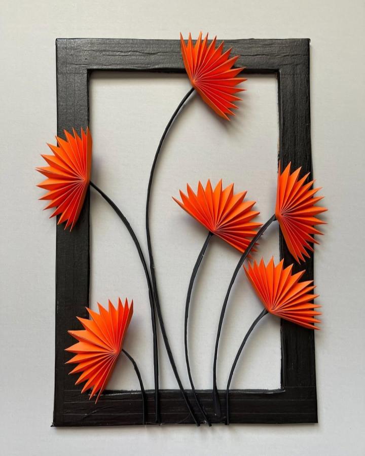 Bright orange accordion-folded paper flowers on black frame stem wall art from @chandanapyla. Modern minimalist craft decor.