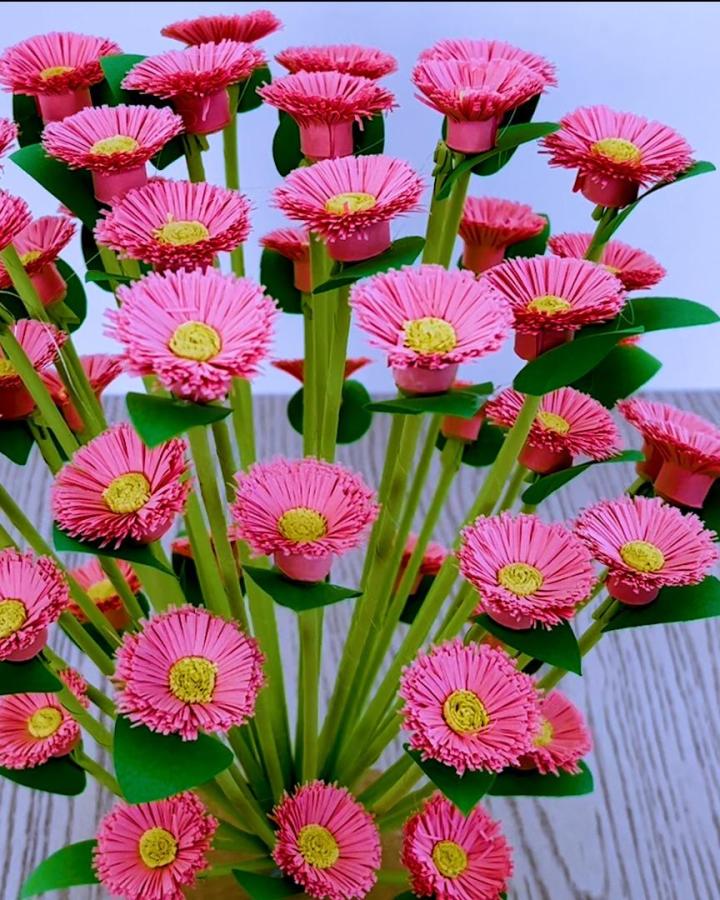 Bright pink fringed paper flowers with yellow centers and green stems by @paper_flowers_artist detailed handcrafted bouquet on wood surface.