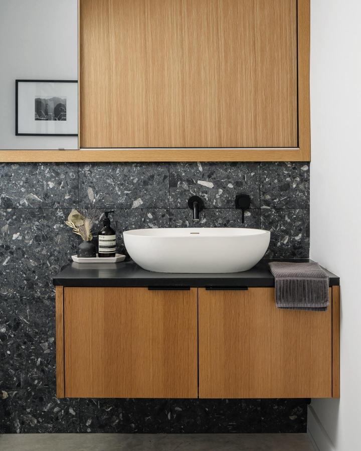 Natural oak floating cabinet with matte black countertop and terrazzo backsplash from @northarrowstudio. Sleek compact basin and hidden storage.