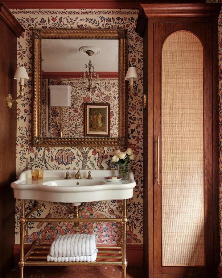 White ceramic sink with vintage brass stand and wooden cane cabinet in a floral wallpapered powder room by @lara_etal. Stylish open and closed storage mix.