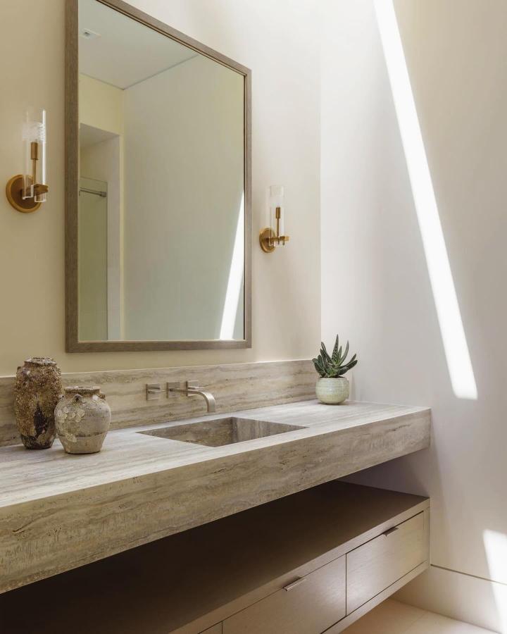 Travertine floating vanity with warm wood accents and brass sconces from @sld_thestudio offers stylish storage in a minimalist powder room.