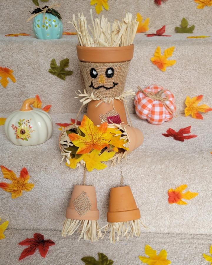 Terracotta pot scarecrow craft with burlap face and raffia hair from @kandf.design sitting among autumn leaves and fabric pumpkins.