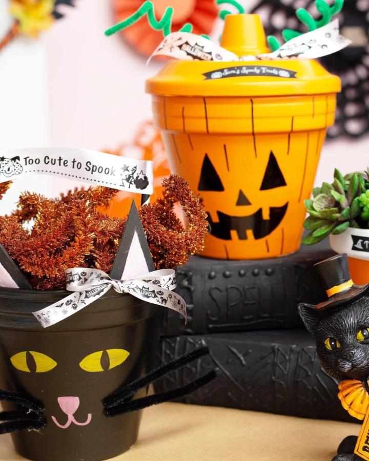 Bright orange and black painted Halloween pots from @creativelivingwithlynnlilly featuring festive jack-o-lantern and cat faces with pipe cleaner accents.