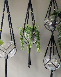Black knotted glass globe plant hangers from @plurque_com. Gold beads cradle trailing greenery against a neutral wall.
