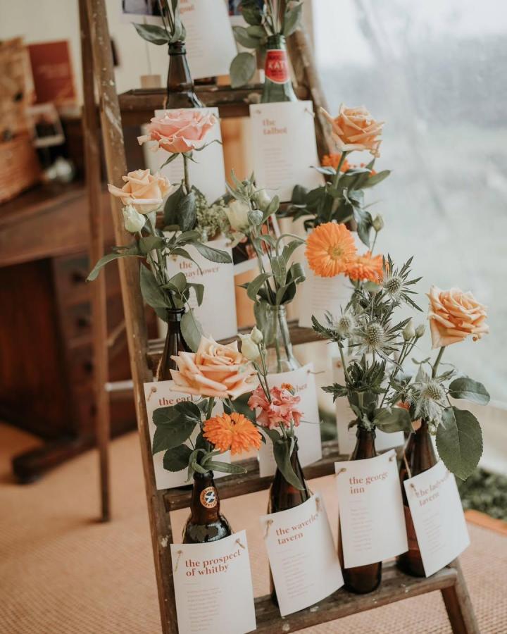 Peach and orange floral bottle display from @abbiewortondesign. Charming diy wedding crafts with cream labels and wooden ladder.