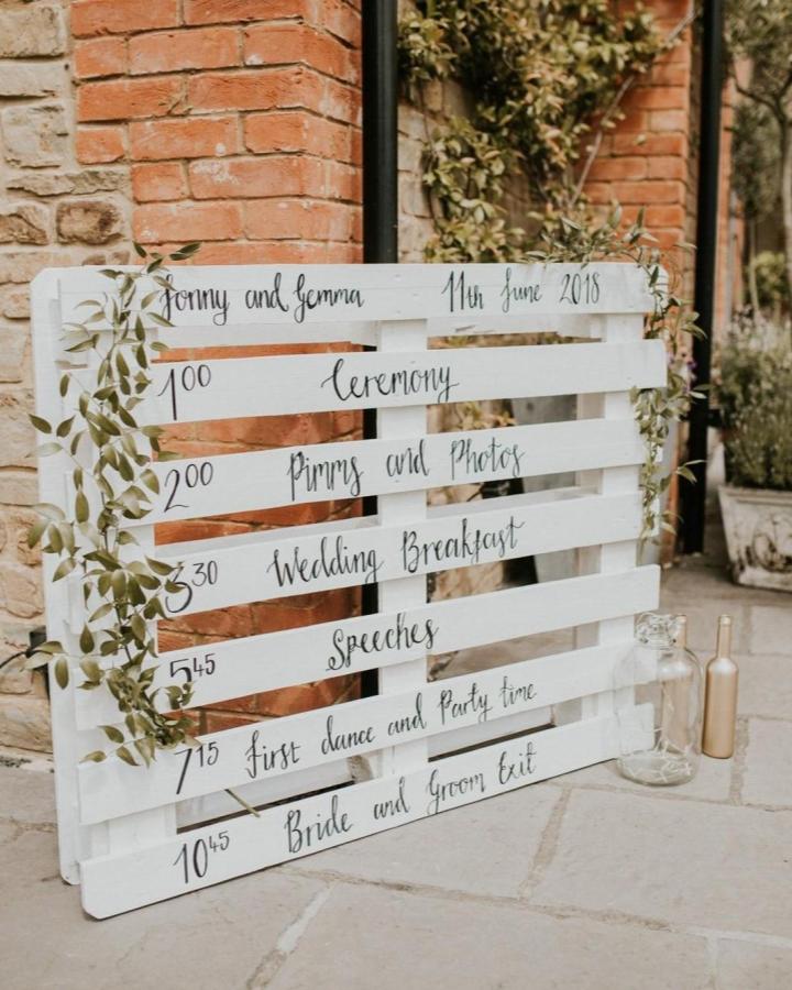 Whitewashed pallet wedding timeline from @rockmywedding. Hand-lettered schedule with trailing greenery.