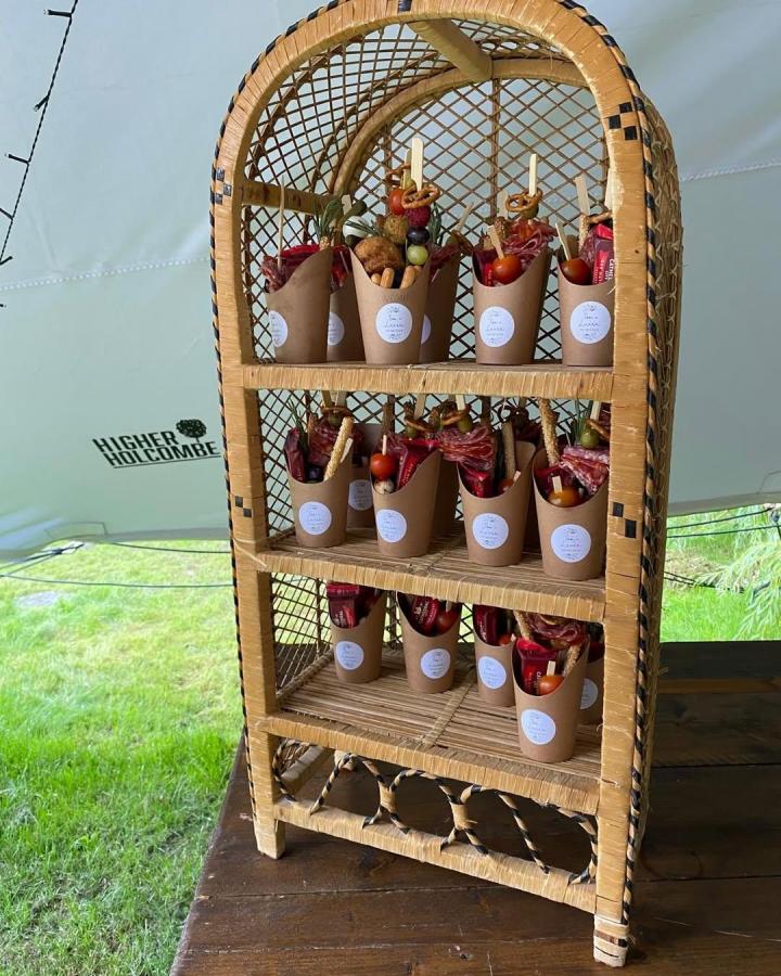 Rustic rattan charcuterie display from @higher.holcombe. Kraft cups hold colorful snacks for a handmade wedding reception.