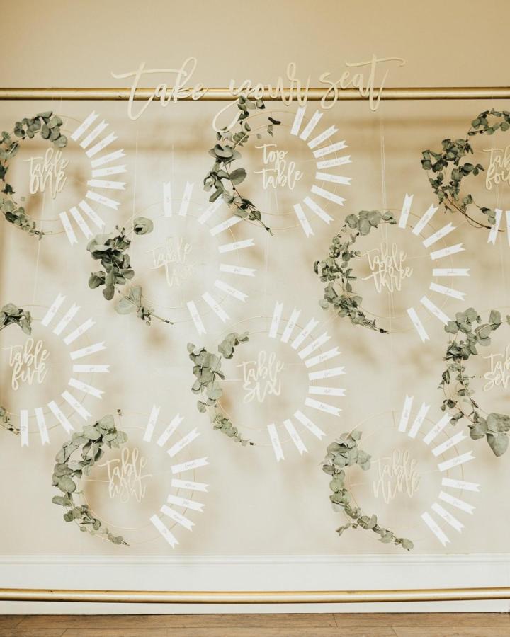 Soft green eucalyptus table-number display from @whimsicalwonderlandweddings. Circular acrylic signs feature white script and sunburst tabs.