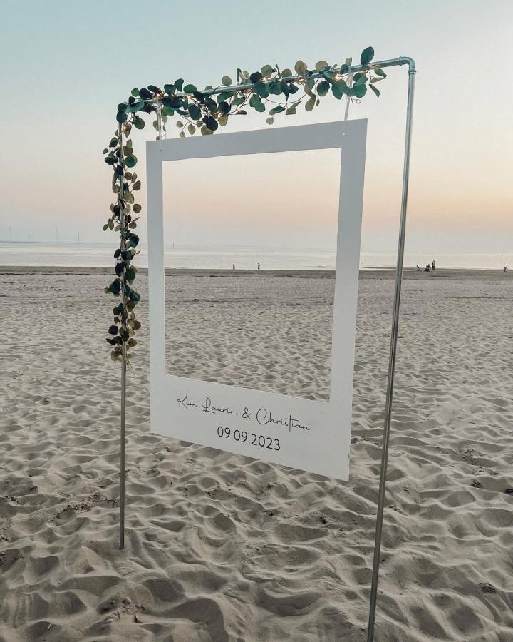 White PVC beach photobooth frame from @kimmlaurine. Eucalyptus greenery and handwritten date add charm to seaside wedding photos.
