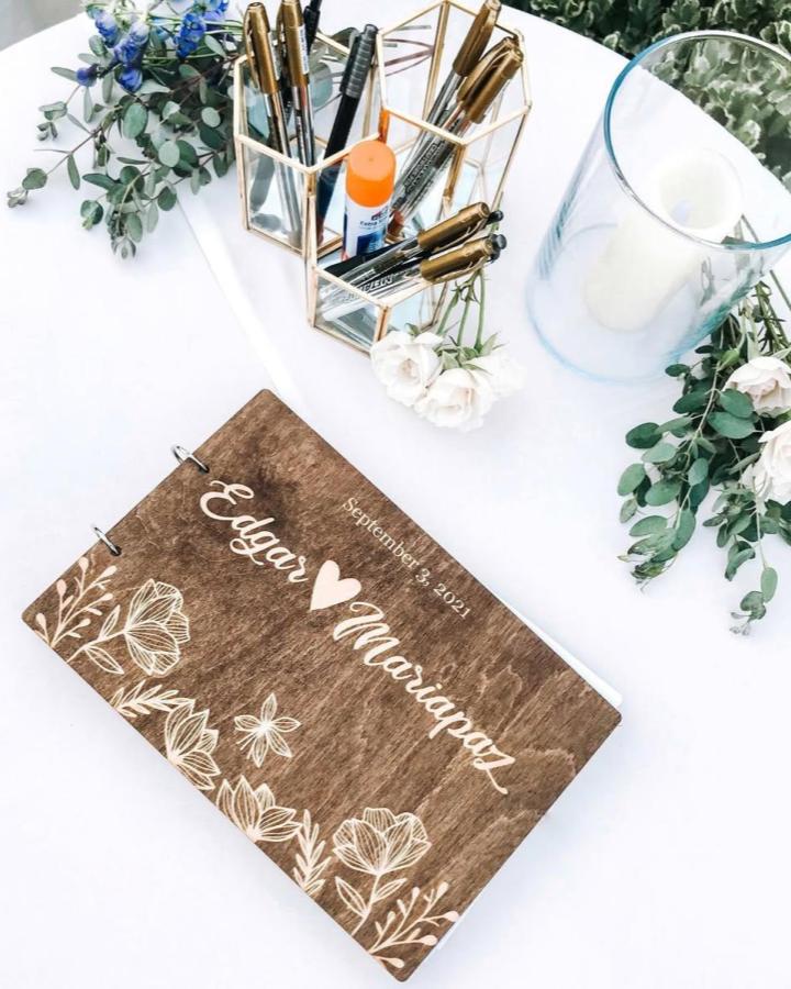 Rustic brown wooden guestbook from @happycamperphotobus. White floral engraving, gold pens, and eucalyptus create a charming wedding keepsake.