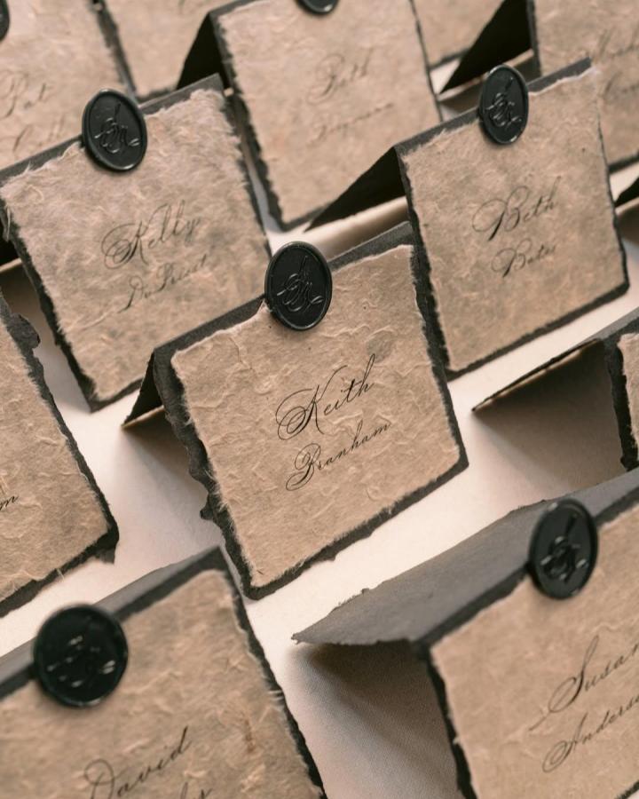 Textured tan folded wedding place cards from @papiradesign. Black wax seals and elegant calligraphy personalize each handmade card.