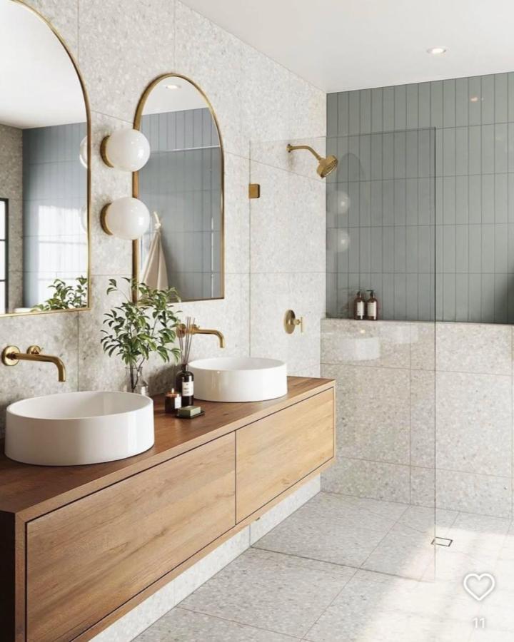 Warm wood double sink bathroom ideas from @innen.architektur. Arched mirrors, white basins, brass fixtures, and gray tile.