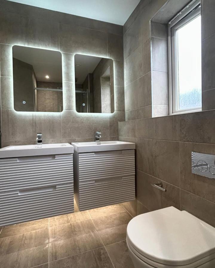 Taupe tiled double vanity bathroom from @custombathroomsglasgow. Backlit mirrors and ribbed white cabinets.