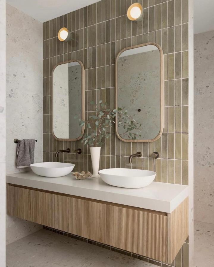 Muted olive-gray tiled double vanity bathroom from @villastyling. Fluted oak cabinetry and rounded mirrors.