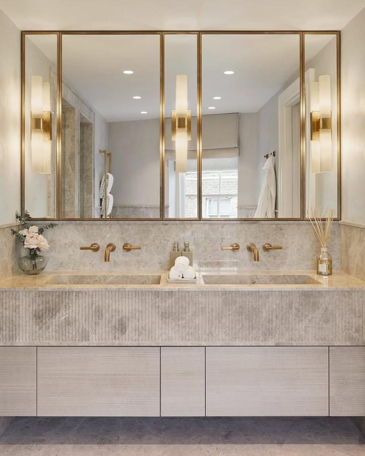 Warm beige fluted double vanity from @conceptbespoke. Gold-framed mirrors, brass faucets, and twin basins.