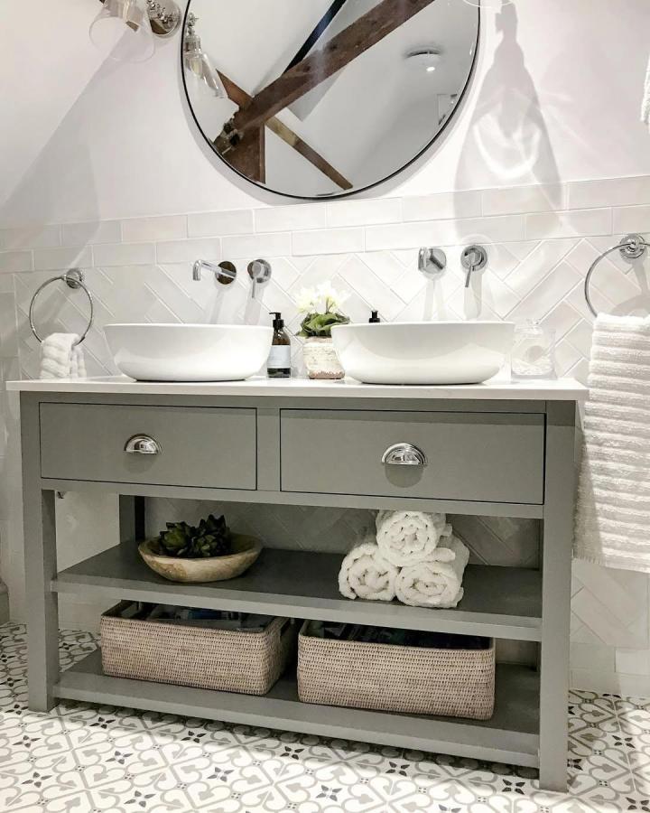 Soft gray double vanity from @our_kentish_barn. White vessel sinks, chevron tile, woven baskets, and patterned flooring.