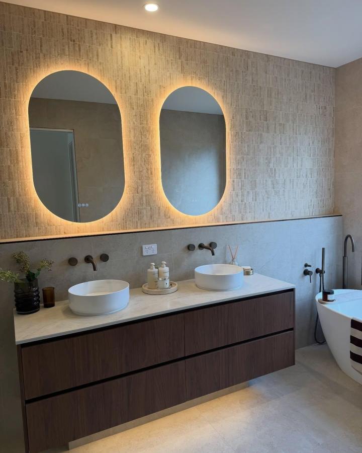 Warm beige double sink bathroom ideas vanity from @thecasaolive. Backlit arched mirrors, white vessel sinks, and dark walnut drawers.
