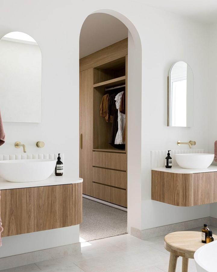 Warm oak fluted double vanity bathroom from @adpaustralia. White vessel sinks, brass taps, arched mirrors, and open storage.