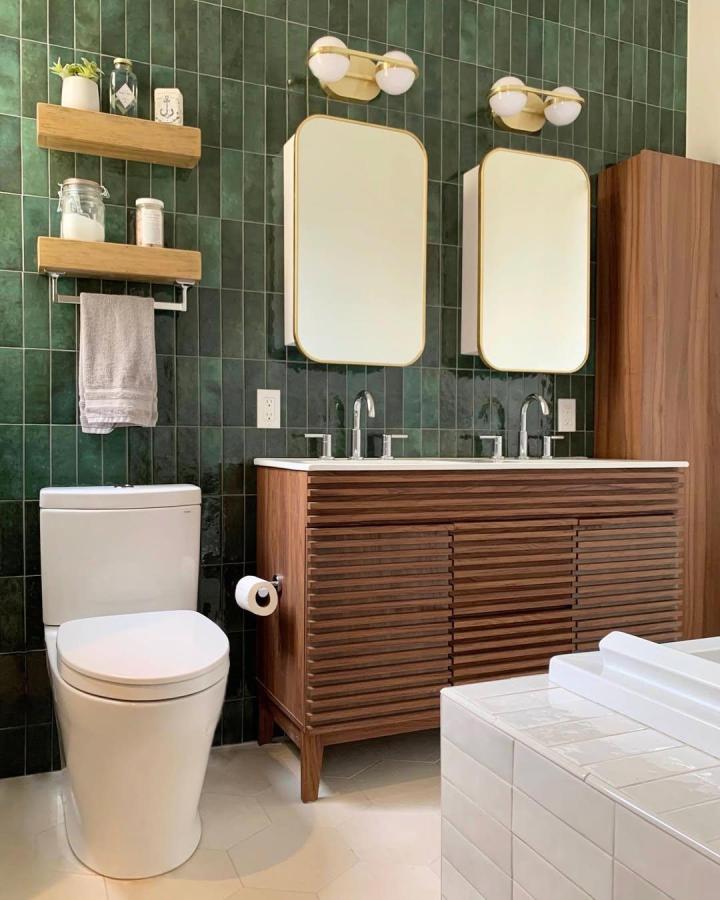 Emerald green tiled double vanity bathroom from @missalicedesigns. Walnut cabinetry, rounded mirrors, and gold globe lights.