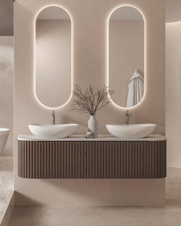 Warm taupe double vanity from @victoria_albert_baths. Arched illuminated mirrors and fluted walnut cabinetry.