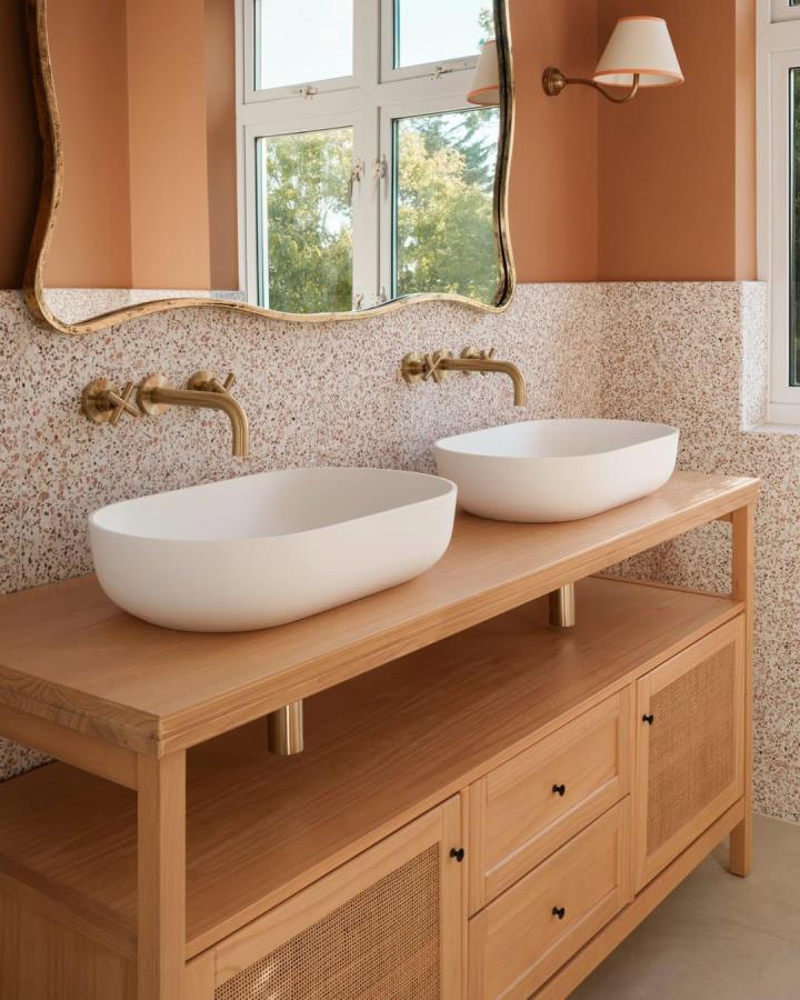 Warm oak double vanity from @aflux_interiors. White vessel basins, brass faucets, terrazzo backsplash, and wavy mirror