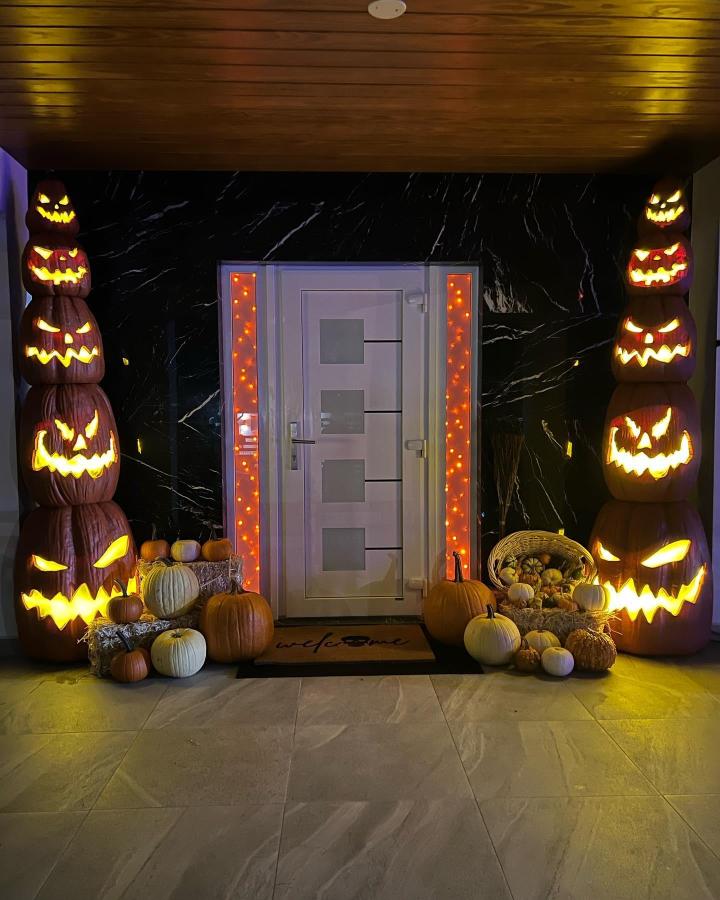 Glowing orange stacked pumpkin towers from @angieanette. Dramatic Halloween entry with gray door, black backdrop, gourds, and warm lights.