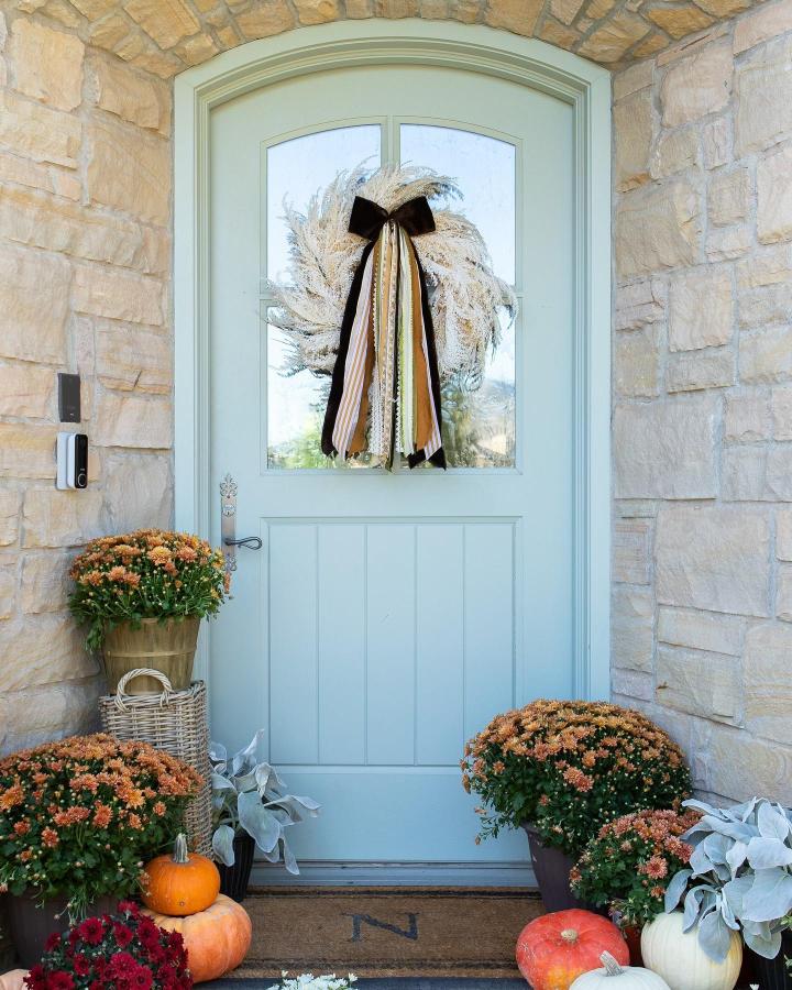 Pale blue fall front door wreath from @.com/havenhaus_co. Cream pampas, black ribbons, orange mums, and pumpkins frame a stone entry.