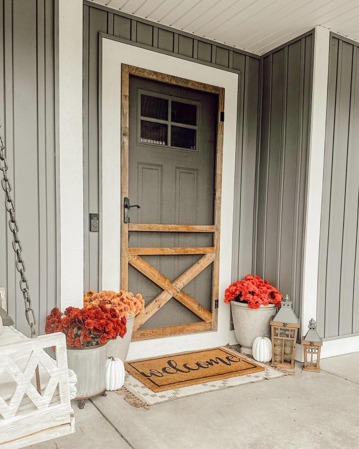 Charcoal-gray farmhouse screen door from @stephaniemartinhome. Orange mums, white pumpkins, lanterns, and layered fall entry mats.