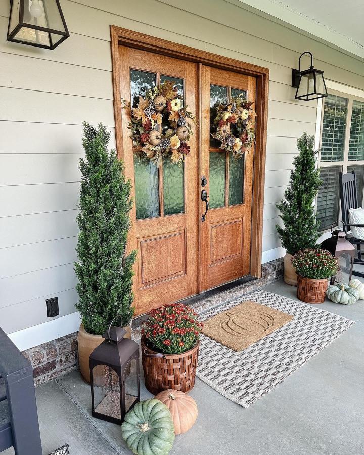 Rust-and-cream floral wreaths on double entry doors from @nearlynaturalfloral. Pumpkins, topiaries, and lanterns frame a cozy autumn porch.