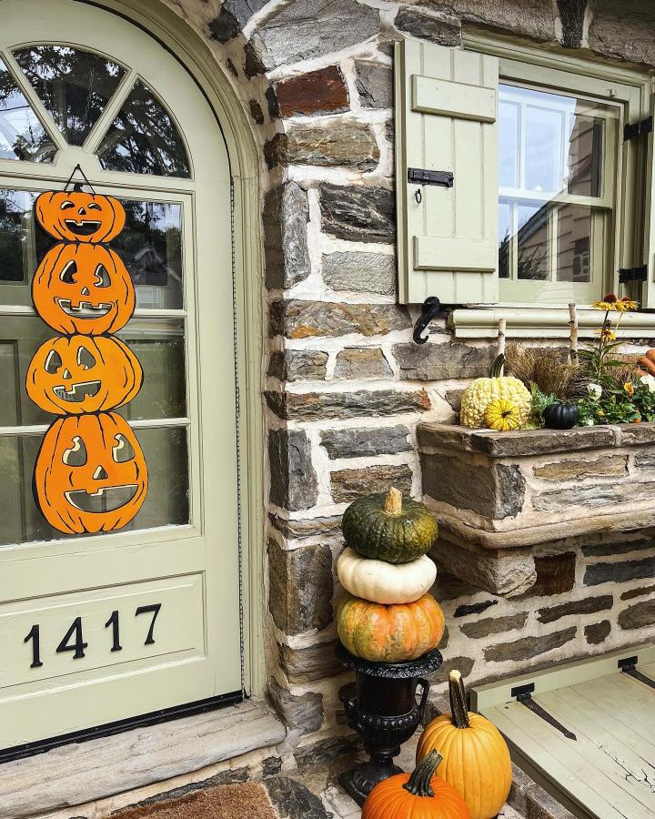 Orange pumpkin door display from @meetmeinphilly. Stacked jack-o’-lanterns and harvest gourds surround a pale green entry.