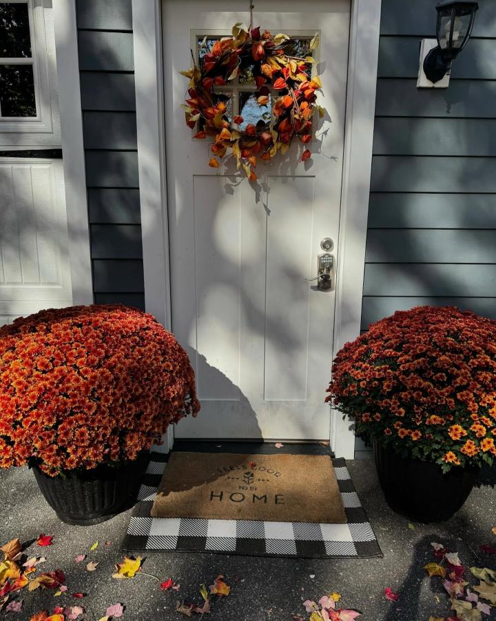 Rust and orange leaf wreath front door from @thegreatpumpkindelivery. Framed by potted chrysanthemums and buffalo-check mats.