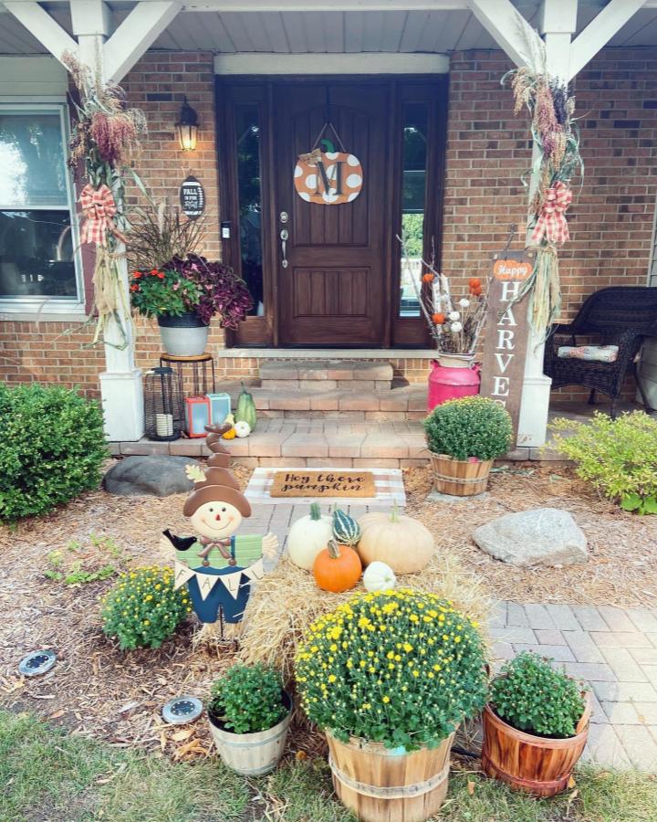 Warm-toned front steps decor from @succulentsandstilettos. Pumpkins, mums, straw, and a cheerful scarecrow welcome guests.