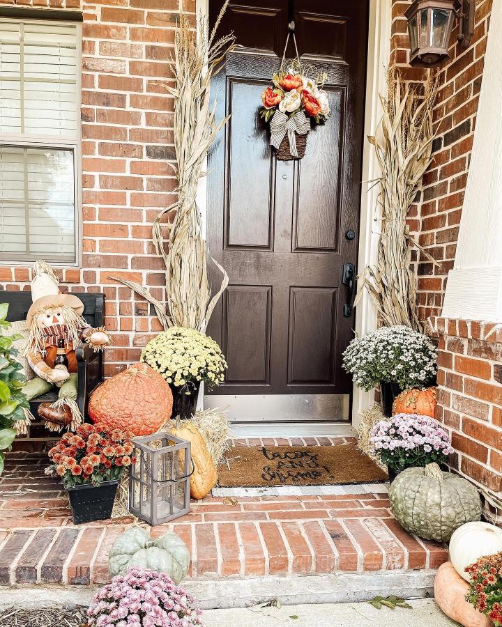 Warm fall front porch from @hometownblessings. Cornstalks, mums, pumpkins, and a harvest wreath frame the dark entry door.