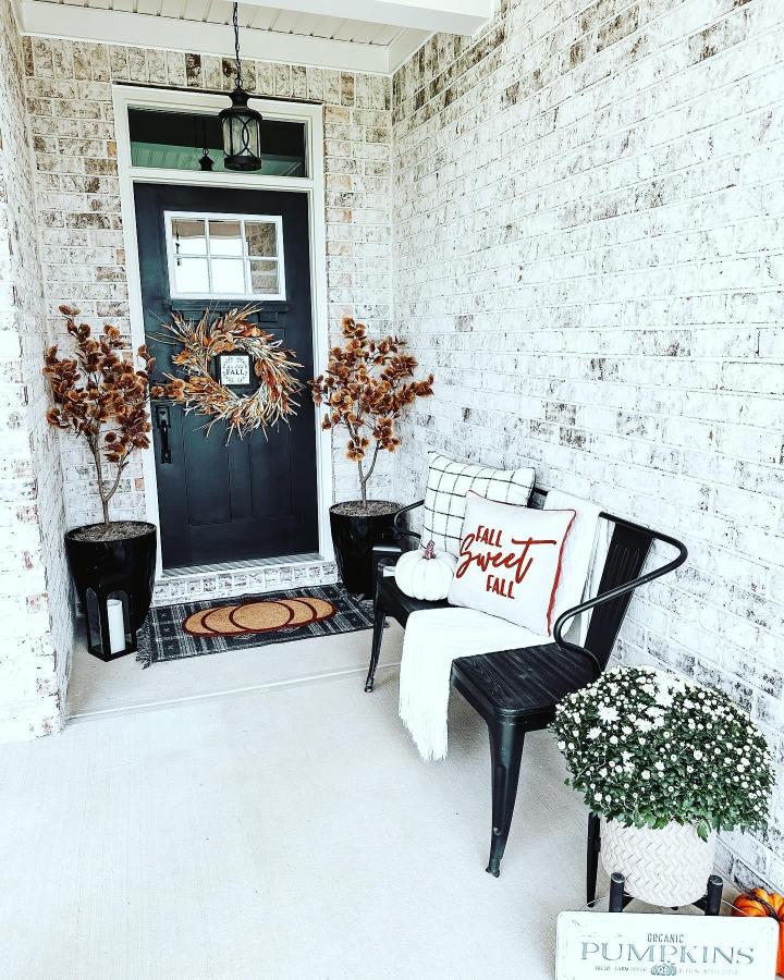 Black door and white brick porch from @the_house_on_boone. Dried stems, wreath, pumpkins, and cozy seating welcome autumn.