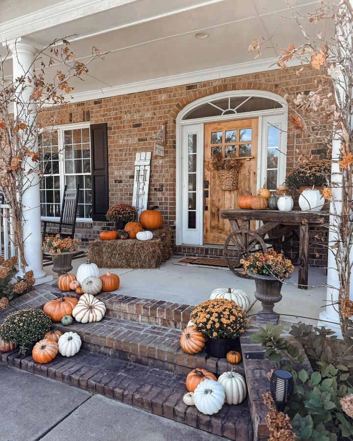 Orange and white pumpkin porch steps from @carolina.cozy. Rustic cart, hay bale, mums, and autumn vines frame the entry.