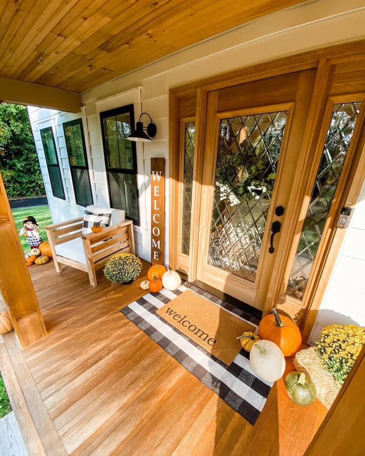 Warm wood porch from @lillianbateman. Buffalo-check runner, pumpkins, mums, and welcoming fall layers surround a glass-paneled door.