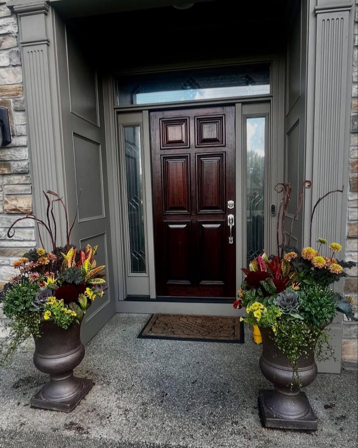 Burgundy and golden floral urn planters from @rootedinjoyplanters frame a dark wood entryway with autumn texture.