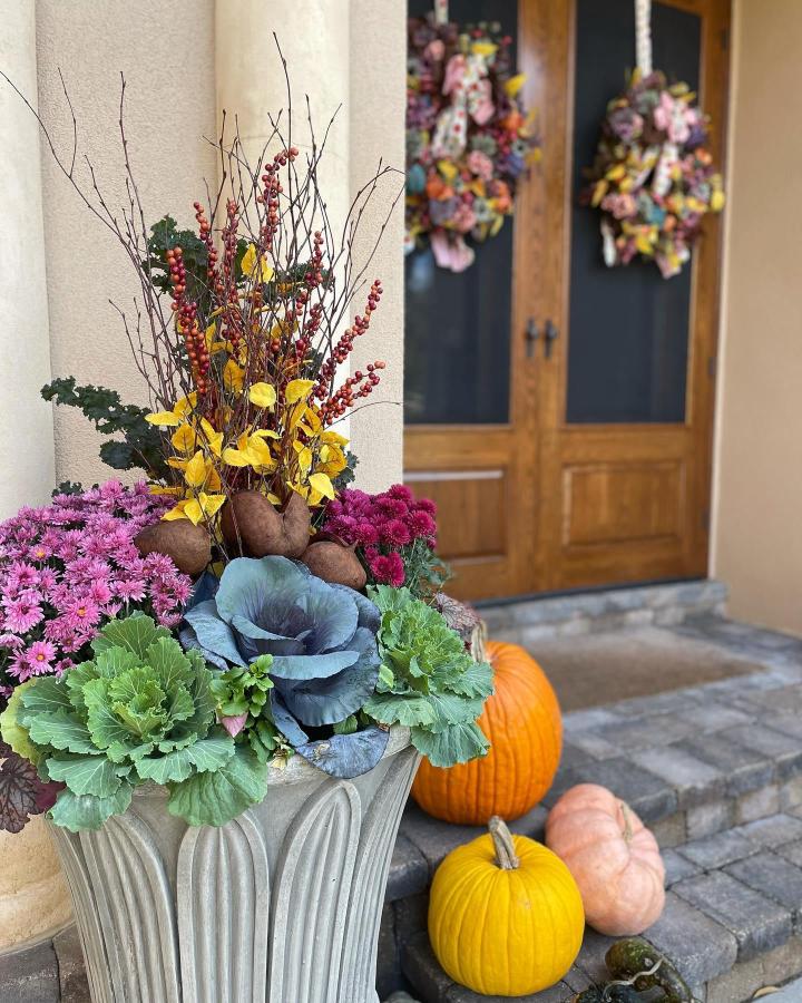 Rustic fall entry planter from @patiopetals. Purple mums, ornamental cabbage, berries, pumpkins, and twig stems fill a fluted urn.