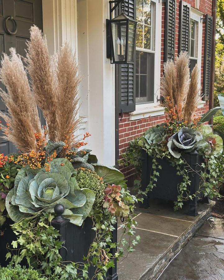 Black layered fall planter ideas from @green.with.ivy. Tall grasses, cabbages, berries, and ivy frame a brick entry.