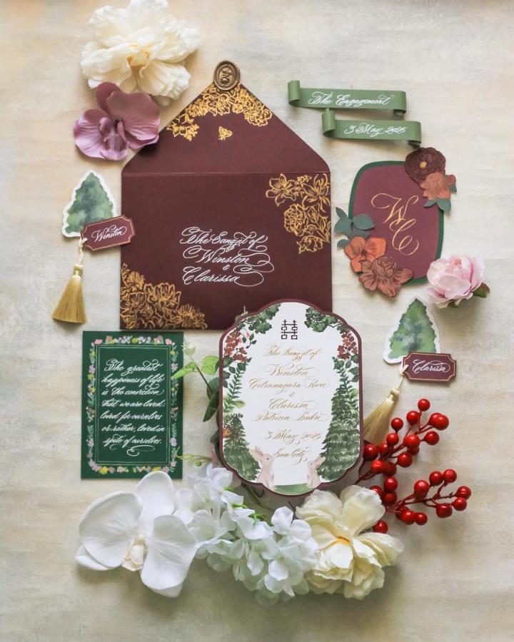 Burgundy and gold calligraphy invitation suite from @calligraphybymercia. Floral details, ribbons, tags, and botanical cards surround the set.