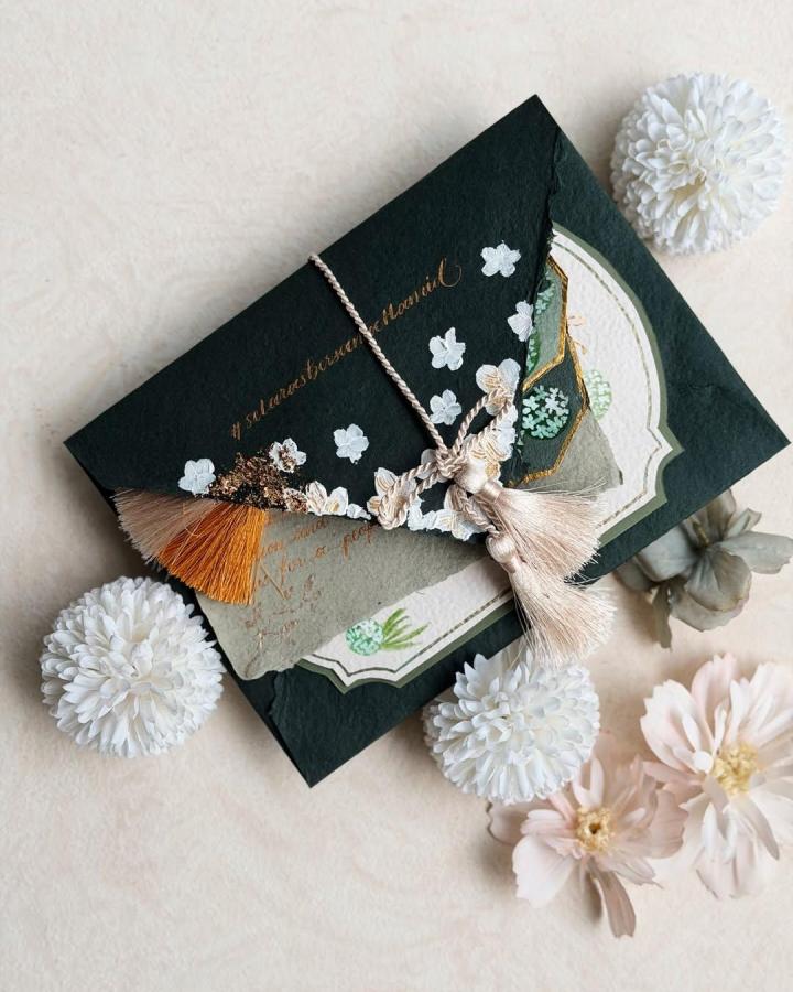 Forest-green tassel invitation from @madebyarti. Handmade wedding stationery with floral accents and gold lettering.