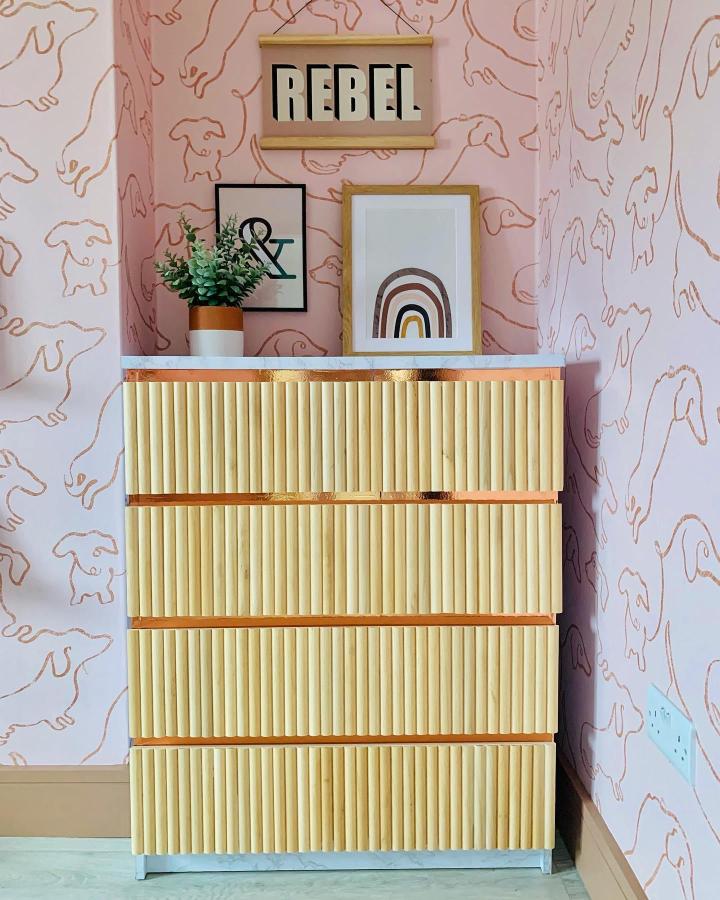 Honey-toned fluted MALM chest from @lovetohomedesign. Pale marble top, gray base, and blush-pink wallpaper create a warm makeover.