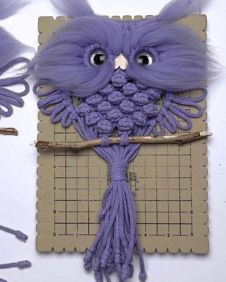 Lavender macrame owl from @cottonkasia. Textured feather knots, cream beak, and twig perch create a whimsical fiber animal.