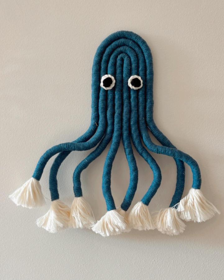 Deep teal macrame octopus wall hanging from @e__macrame. Cream tassels and googly eyes decorate curling tentacles.