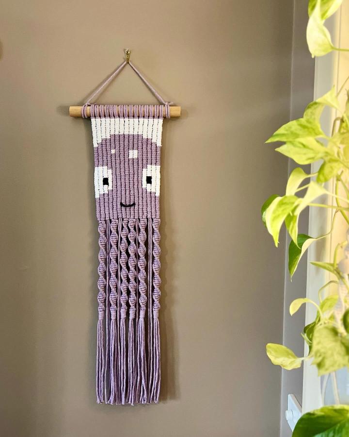 Lavender cat wall hanging from @macramecollection_. Knotted cotton face with twisted fringe beside a leafy plant.