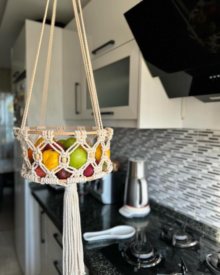 Cream knotted hanging fruit basket from @alacatiartmacrame. Wooden rail and tassel hold colorful produce in a bright kitchen.