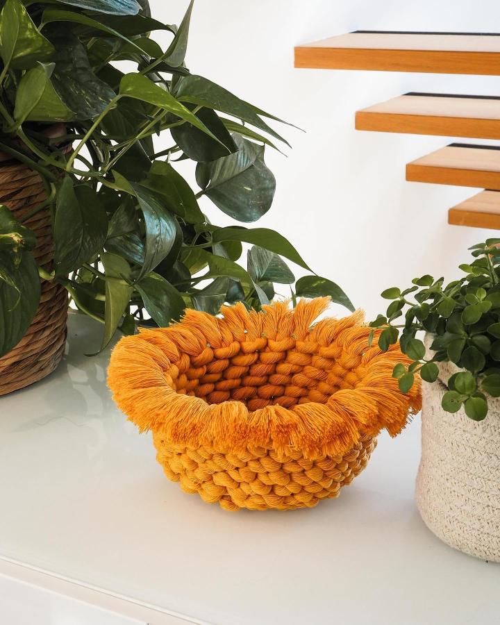 Orange fringed woven basket from @macramemakersclub. Chunky knots beside leafy houseplants.