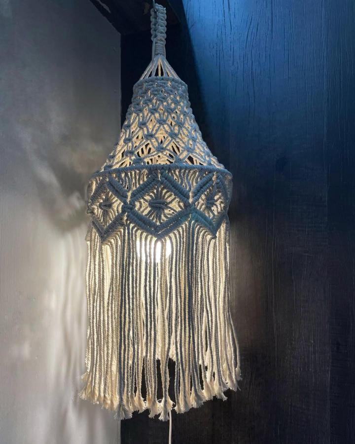 Ivory macrame pendant light from @themacthings. Intricate knots and long fringe glow against a dark blue wall.
