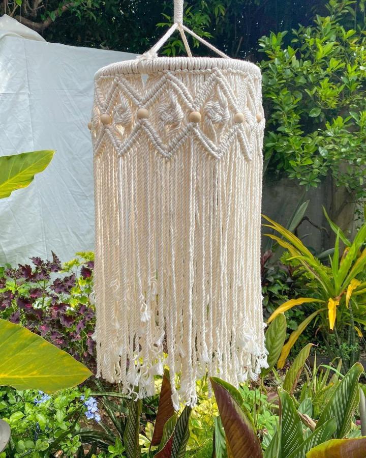 Ivory cotton macrame chandelier from @khitemacrame. Zigzag knots, wooden beads, and long fringe hang among lush garden greenery.