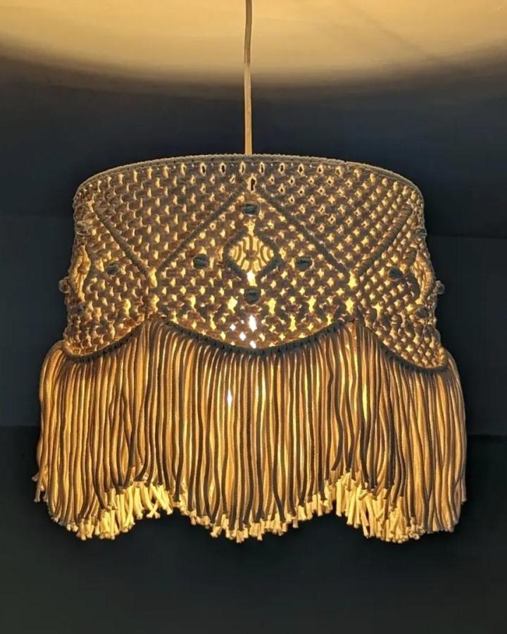 Golden macrame chandelier from @quiet.trace.studio. Woven shade with dramatic fringe glows against a dark wall.